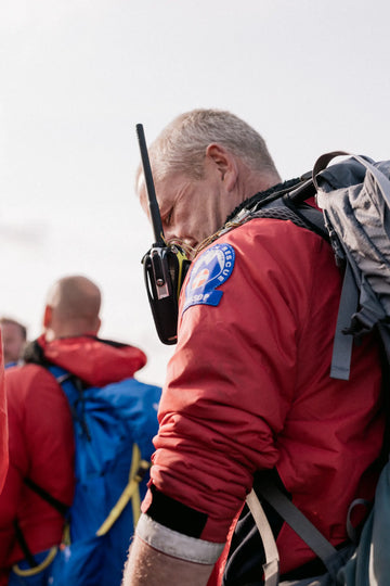 Person wearing a red jacket with a walkie-talkie and backpack, looking down. Buffalo Systems Mountain Rescue