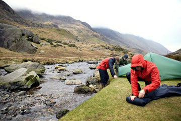 Two people setting up a tent by a stream in a mountainous area