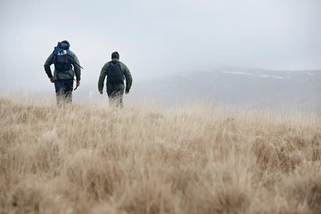 Two people walking through tall brown grass on a foggy day.