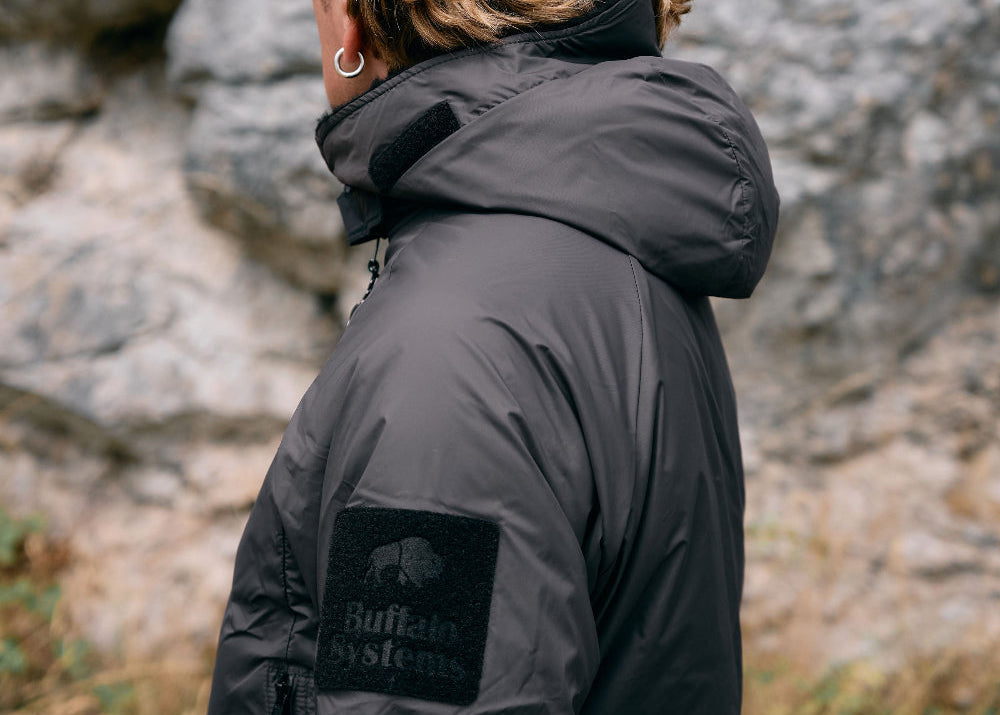 A person with blonde hair seen from the back wearing a black hooded puffer jacket with a 'Buffalo Systems' logo patch on the sleeve, standing outdoors against a rocky cliff face.