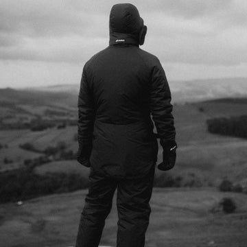 Person wearing a black jacket with a hood, standing in a landscape.